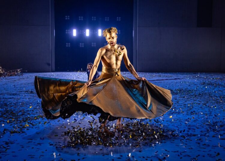 A man on stage painted all gold with a gold leaf crown and large gold skirt flowing outwards like he's been twirling around.