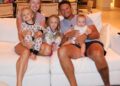 Brittany and Patrick Mahomes sit together on a white couch at home with their three children, Sterling, Bronze, and baby Golden, all smiling in their pajamas for a cozy family photo.