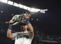 A Seattle Mariners baseball player lifts a white guitar with black accents up in the air in a stadium