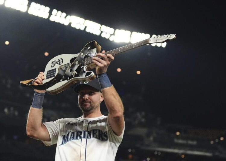 A Seattle Mariners baseball player lifts a white guitar with black accents up in the air in a stadium