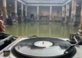 BBC A deck with a vinyl playing is in the foreground. In the background is a Roman thermal bath, which looks like an open-top green rectangular pool, with stone pillars surrounding it. There are people sitting around the pool.