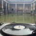 BBC A deck with a vinyl playing is in the foreground. In the background is a Roman thermal bath, which looks like an open-top green rectangular pool, with stone pillars surrounding it. There are people sitting around the pool.