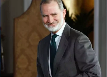 A bearded man in a grey suit smiles as he stands in a stylish interior setting.