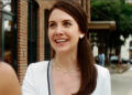 Alison Brie, wearing white and smiling big as Sydney Prescott's publicist Rebecca Walters in Scream 4.
