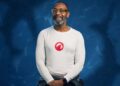 Sir Lenny Henry pictured in front of a dark blue background, smiling ahead. He is wearing a long sleeved white t-shirt with a red emblem on the front.