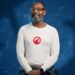 Sir Lenny Henry pictured in front of a dark blue background, smiling ahead. He is wearing a long sleeved white t-shirt with a red emblem on the front.