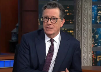 Stephen Colbert wearing a suit and tie while sitting at a desk hosting The Late Show with Stephen Colbert. .