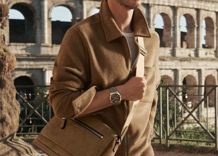 a male figure stands near the colosseum wearing a brown jacket and holding a brown bag