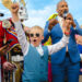 A lively crowd with a young boy in sunglasses and a blue vest raising his arms. A man in a blue suit holds a microphone. People cheer around them.