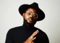 A man wearing a black hat, round glasses, and a black turtleneck poses against a plain background, with his hands clasped together and rings on his fingers.