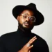 A man wearing a black hat, round glasses, and a black turtleneck poses against a plain background, with his hands clasped together and rings on his fingers.