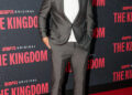 Travis Kelce smiles on the red carpet in a gray suit over a white T-shirt, paired with white sneakers and dark sunglasses. The backdrop features the ESPN Original series The Kingdom.