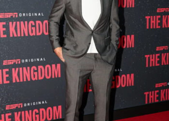 Travis Kelce smiles on the red carpet in a gray suit over a white T-shirt, paired with white sneakers and dark sunglasses. The backdrop features the ESPN Original series The Kingdom.