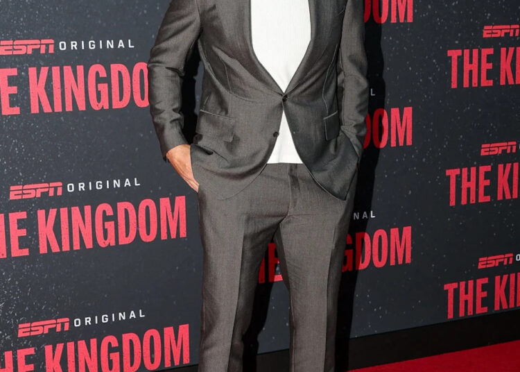 Travis Kelce smiles on the red carpet in a gray suit over a white T-shirt, paired with white sneakers and dark sunglasses. The backdrop features the ESPN Original series The Kingdom.
