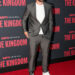 Travis Kelce smiles on the red carpet in a gray suit over a white T-shirt, paired with white sneakers and dark sunglasses. The backdrop features the ESPN Original series The Kingdom.
