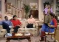 A still frame from an episode of The Cosby Show showing the inside of a home, with a large staircase in the background, with man, a woman and a young woman sitting on a couch, a young man standing to the right of the couch and to the left of a young woman sitting in a chair.