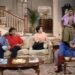 A still frame from an episode of The Cosby Show showing the inside of a home, with a large staircase in the background, with man, a woman and a young woman sitting on a couch, a young man standing to the right of the couch and to the left of a young woman sitting in a chair.