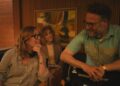 A woman in a striped shirt with headphones around her neck looks at a man leaning on a chair as another woman looks on.