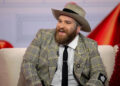 Teddy Swims smiles in a plaid suit jacket and hat.