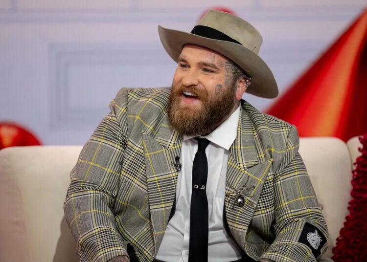 Teddy Swims smiles in a plaid suit jacket and hat.