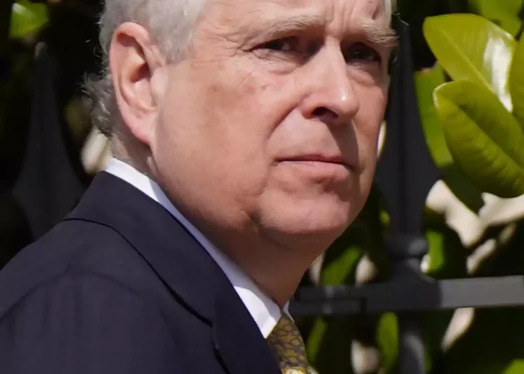 Prince Andrew in a dark suit and yellow tie, in profile, outdoors with green leaves in the background
