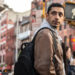 Riz Ahmed with a backpack near a crosswalk in Relay.