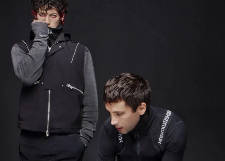 Tyler Joseph and Josh Dun pose against a black backdrop dressed in dark outfits