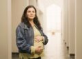 A woman cradling her baby bump as she smiles and looks ahead.