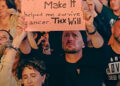 A woman's headband seems to go over the wrist of the woman in front, while her own hand morphs into the man holding up a sign (YouTube/Will Smith)