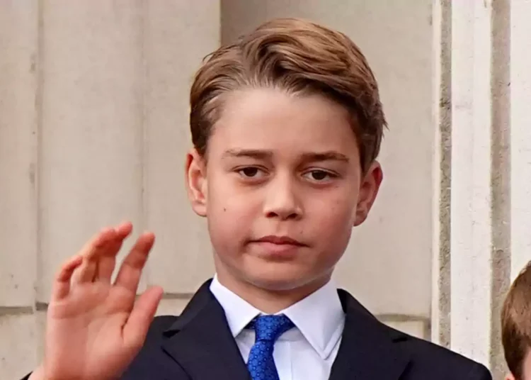 Prince George in a dark suit and blue tie raises his hand in front of a stone building.