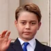 Prince George in a dark suit and blue tie raises his hand in front of a stone building.