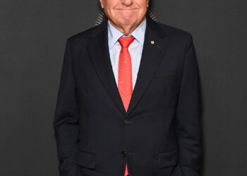 Lorne Michaels stands on the red carpet wearing a navy suit with a light blue dress shirt and red tie, hands in his pockets, smiling at the camera.