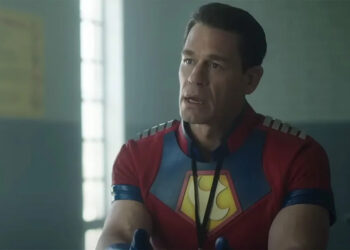 John Cena with a visitor badge on sitting at a prison table in Peacemaker Season 2.