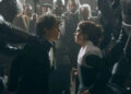 Two actors in period costumes face each other intensely amidst armored figures, while onlookers watch. Scene suggests dramatic tension