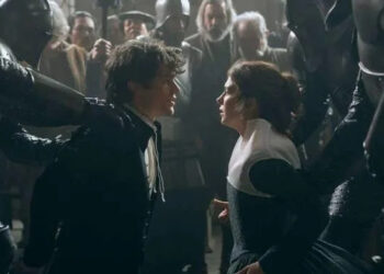 Two actors in period costumes face each other intensely amidst armored figures, while onlookers watch. Scene suggests dramatic tension
