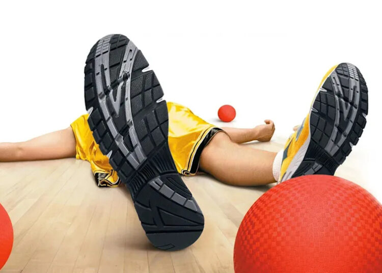 Person in yellow shorts lying on a gym floor, surrounded by red dodgeballs, with sneakers prominently visible in the foreground.