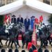 Rows of soldiers riding black horses pass by four people standing in a white tent