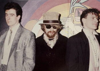Joe Hughes (right), Dave Stewart and David Freeman, standing by a wall. Joe is looking to the camera, Dave Stewart, with glasses and a hat on, also to the camera and David to the right. A mural is behind them.