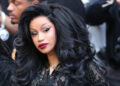 Cardi B with voluminous black hair and glamorous makeup, wearing a black outfit, surrounded by photographers.