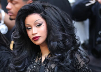 Cardi B with voluminous black hair and glamorous makeup, wearing a black outfit, surrounded by photographers.