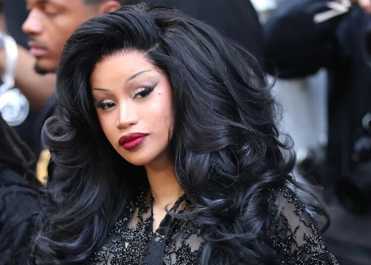 Cardi B with voluminous black hair and glamorous makeup, wearing a black outfit, surrounded by photographers.