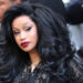 Cardi B with voluminous black hair and glamorous makeup, wearing a black outfit, surrounded by photographers.