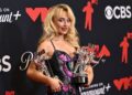 a woman with blonde hair in a sparkling dress holds three trophies in her arms as she smiles at the camera on a red carpet for an awards event