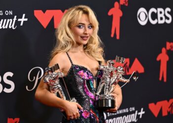 a woman with blonde hair in a sparkling dress holds three trophies in her arms as she smiles at the camera on a red carpet for an awards event
