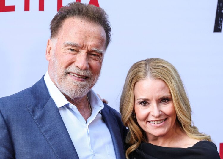 Arnold Schwarzenegger smiling on the red carpet with girlfriend Heather Milligan, both dressed elegantly