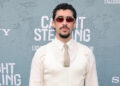 A man with curly hair and sunglasses stands in front of a backdrop with text, wearing a white shirt, tie, and vest.