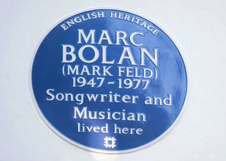 A close up of a blue English Heritage plaque on a white wall.