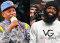 Two men side by side; one in a sports jacket and cap speaking into a microphone, the other in a white shirt smiling.