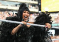 A woman in a black feathered outfit holds a microphone in a wrestling ring, smiling energetically at an event with a large crowd.