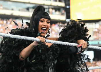 A woman in a black feathered outfit holds a microphone in a wrestling ring, smiling energetically at an event with a large crowd.